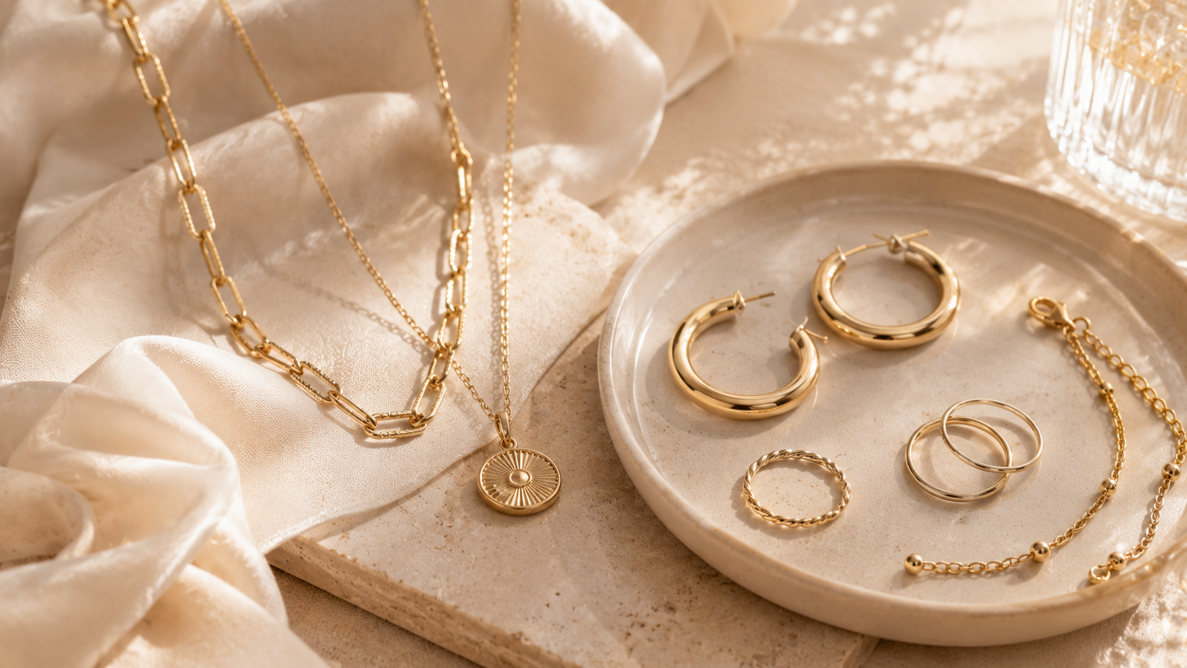 Minimalist gold jewelry including layered chain necklaces, hoop earrings, rings, and a delicate bracelet arranged on soft fabric and ceramic trays under warm natural sunlight.